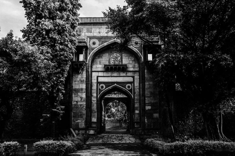 In monochrome, the garden gate frames a smaller arch beyond it; branches and deep shade enclose the path to the tomb.