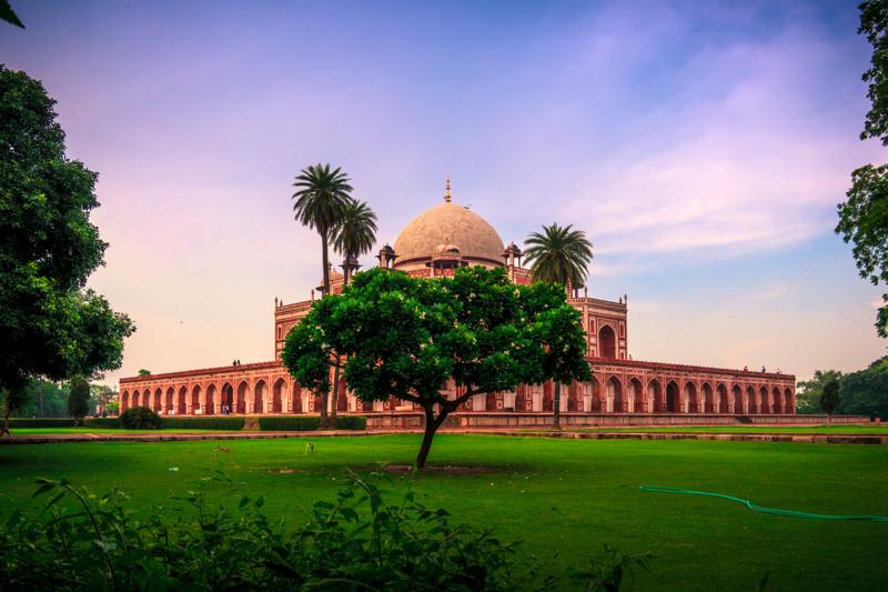 A palm and leafy tree partly screen Humayun’s Tomb, its dome and long red façade set beneath a clear pastel sky.
