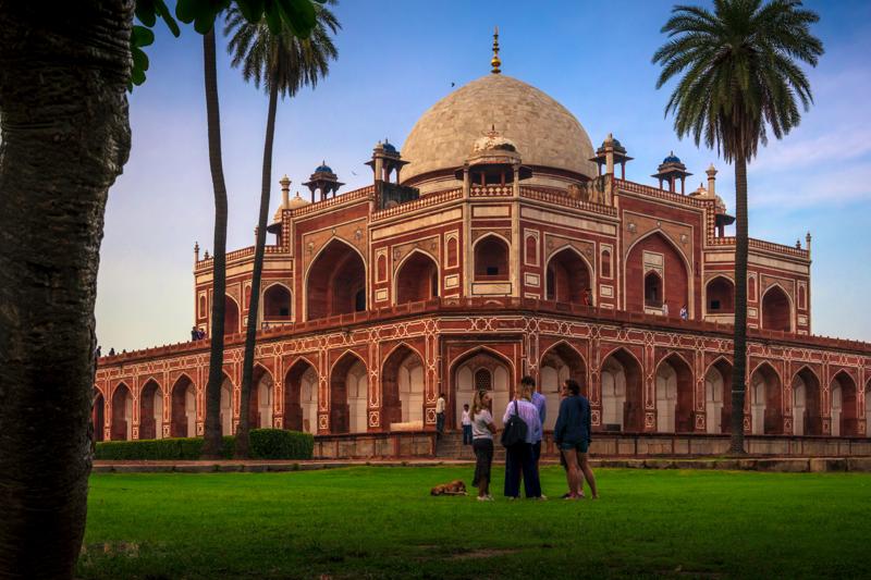 Humayun’s Tomb stands between tall palms, visitors crossing the broad lawn below its red arches under a pale blue evening sky.