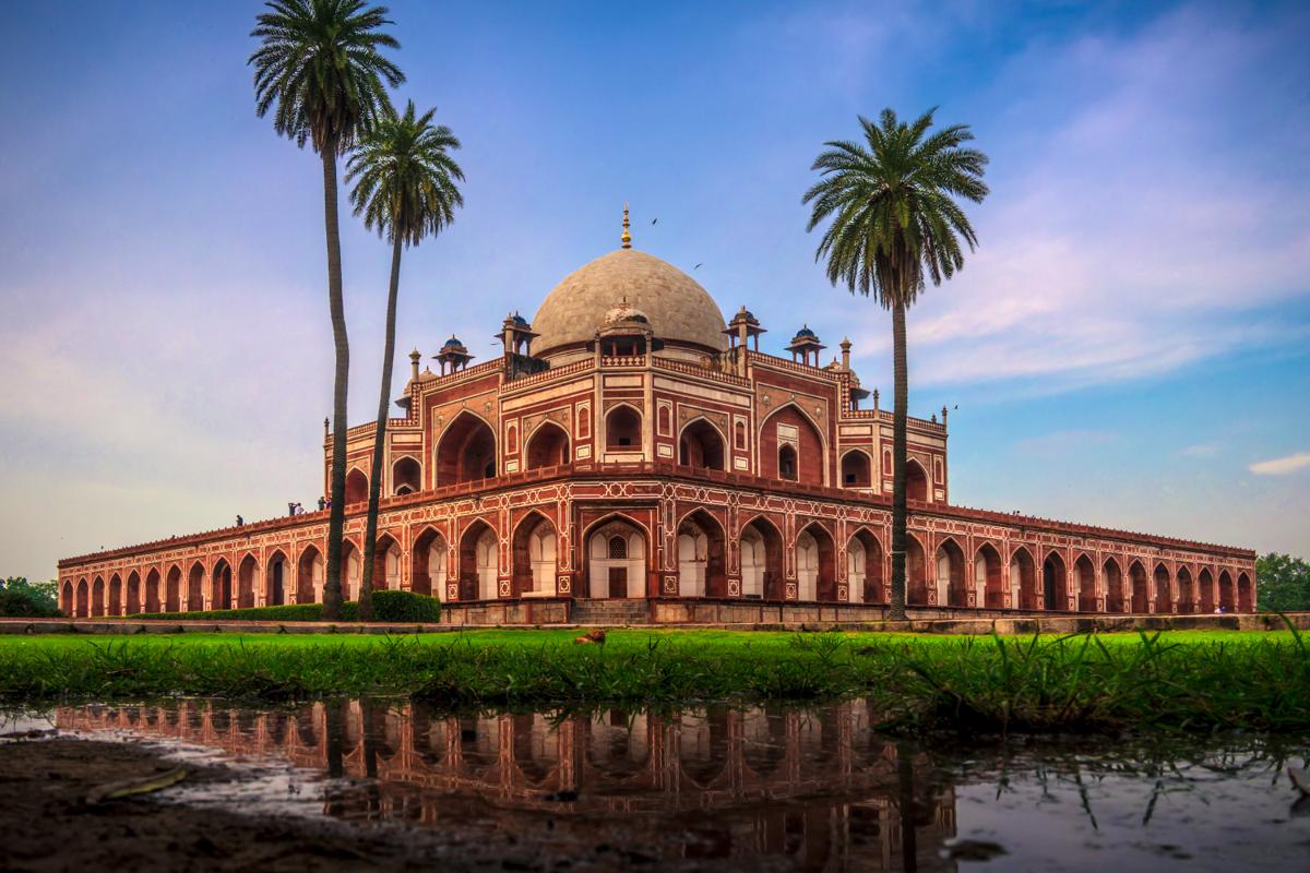 The red sandstone tomb appears upside down in the pool, its arches and palms softened by ripples across the foreground water.