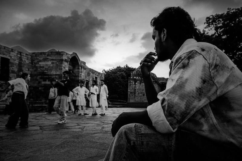 An older man rests on the pavement before the Qutub complex, his dark outline separated from the bright stone and distant visitors.