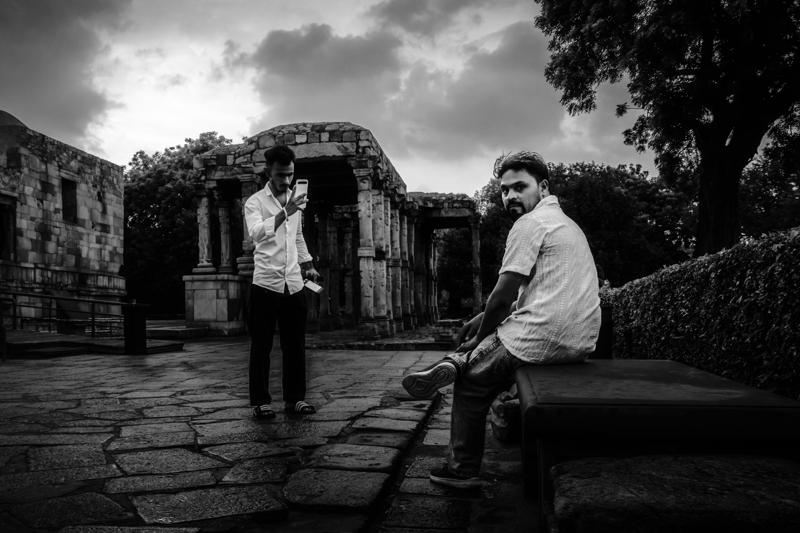 Several visitors cross the stone courtyard beneath Qutub Minar, the tall tower rising beyond the dark silhouettes and ruined walls.