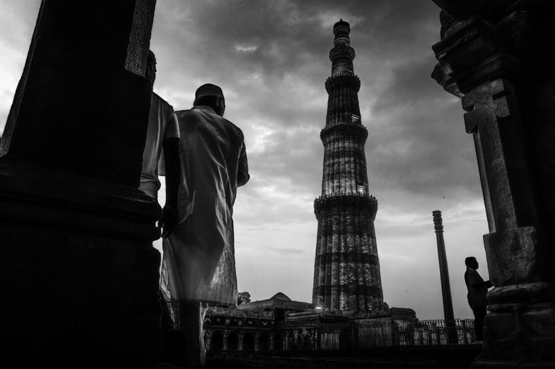 A standing figure looks up from the shadowed courtyard while Qutub Minar climbs through a bright, clouded monochrome sky.