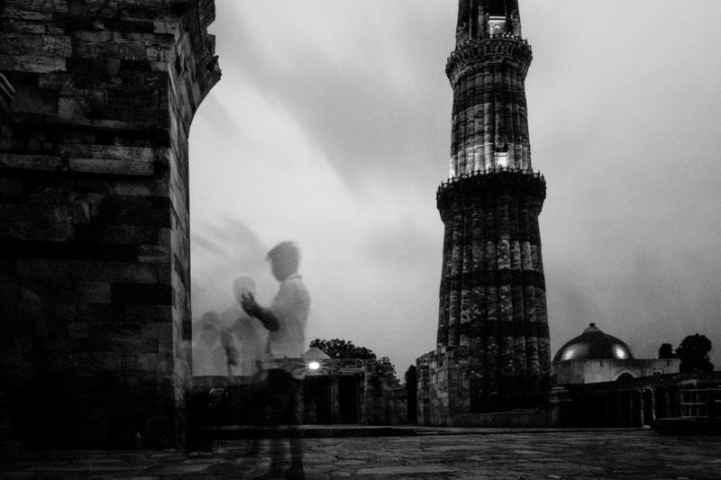 The lit minaret and shaded gateway appear in a shallow puddle, their monochrome reflection broken by the blurred figure crossing the courtyard.