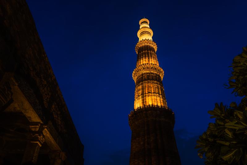 Qutub Minar glows through a narrow opening in the old architecture, its amber shaft set against the cloudless dark blue.