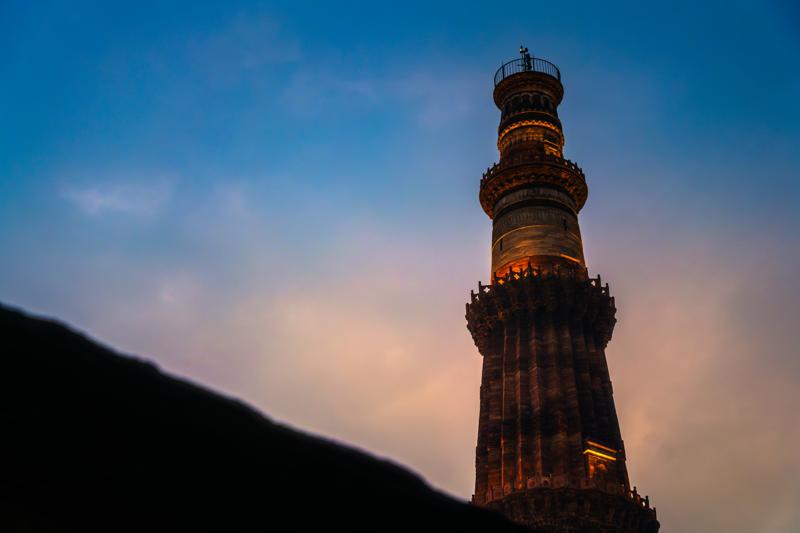 The lit tower rises alone against a clear evening sky, its warm stone separated from the dark slope cutting across the foreground.