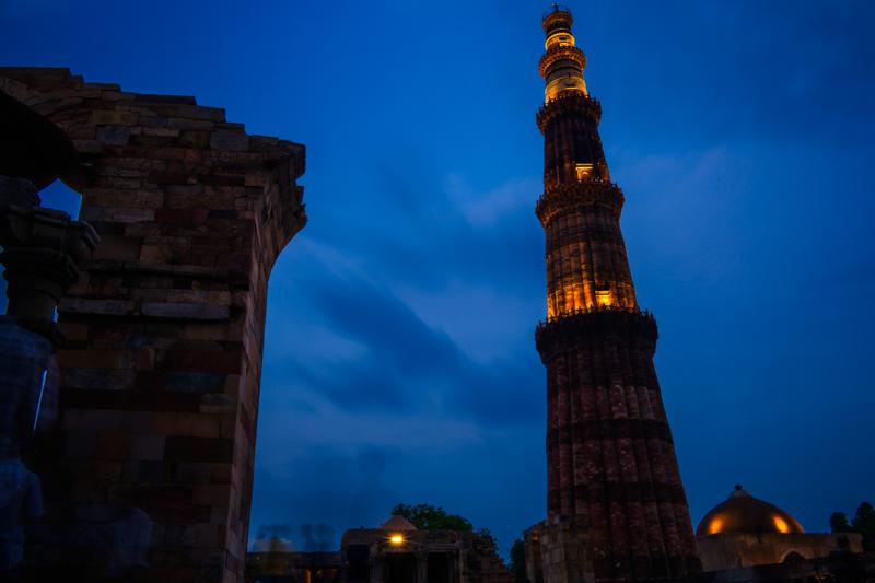 The lit tower stands beyond old stone walls, its amber details rising sharply against the deep blue evening sky.