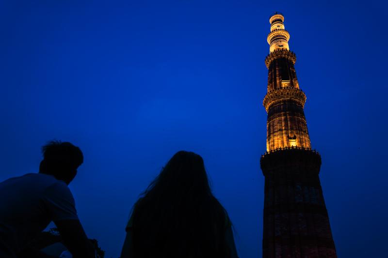 The illuminated Qutub Minar rises above two dark foreground figures, its warm stone sharply outlined against the blue evening sky at dusk.