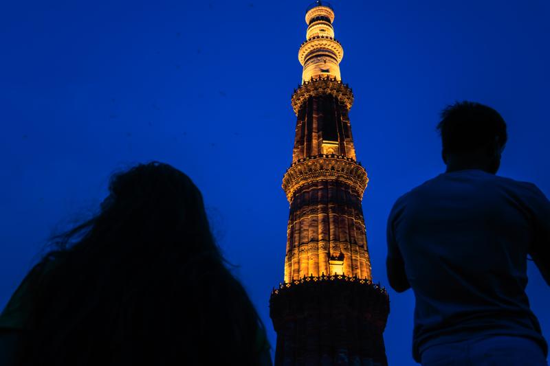 Two figures look up at the illuminated tower, their heads reduced to silhouettes against its amber stone and the deep blue sky.