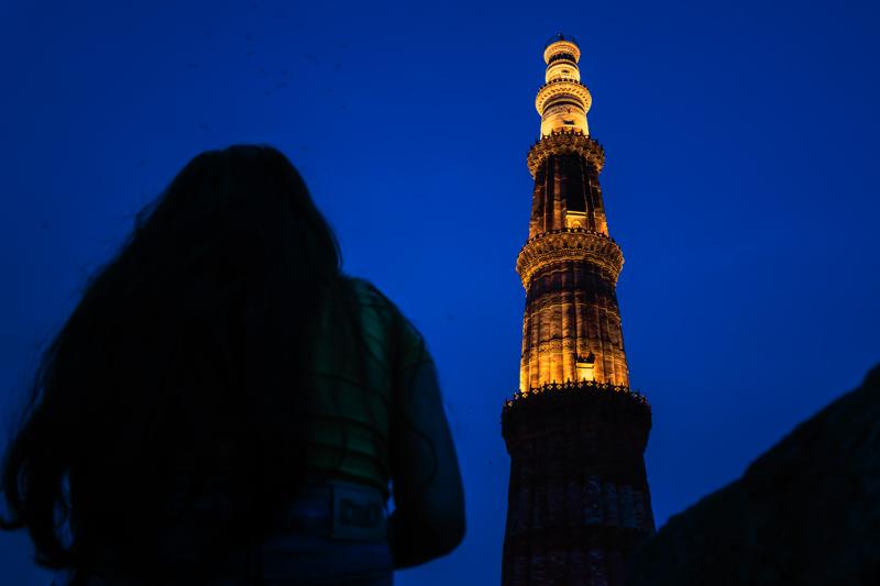 A passerby crosses the dark gateway while the lit minaret stands in the distance, the heavy stone opening framing the pale courtyard.