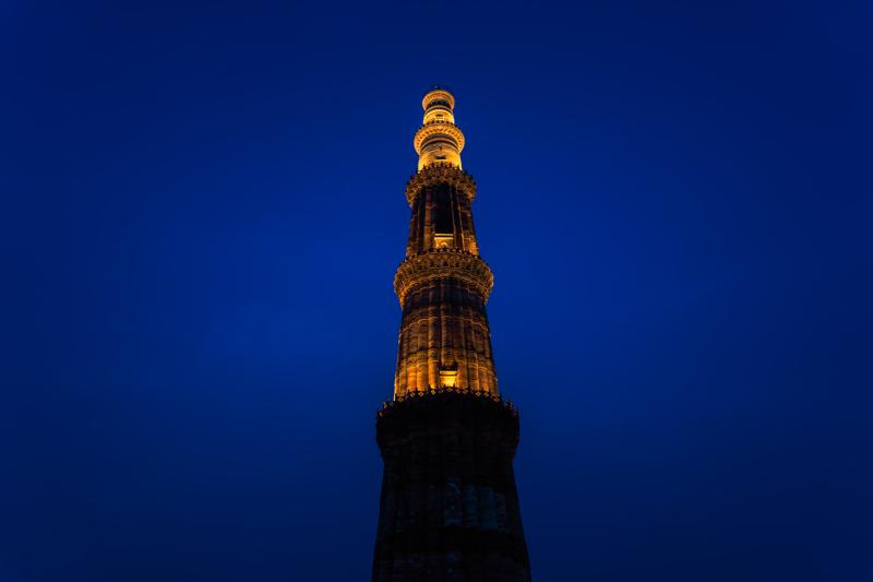 Qutub Minar rises cleanly against the saturated evening sky, its lit stone narrowing to a point above the dark ground.