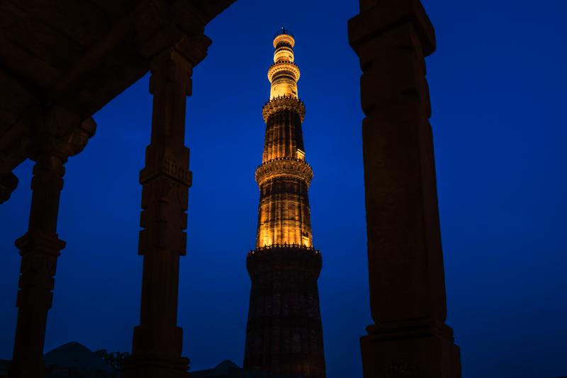 The lit minaret stands between heavy stone columns, with a blurred visitor in the foreground adding scale to the carved passage.