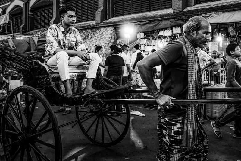 A seated passenger rides past shopfronts as a man walks beside the handcart, with stacked goods and striped awnings crowding the street.