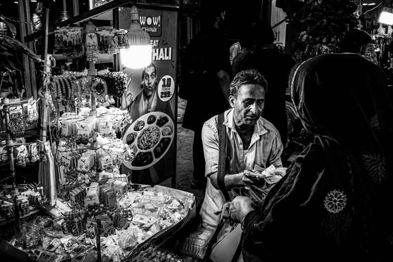 The shopkeeper sits beneath a dark signboard, his face picked out among hanging packets and stacked containers in the narrow stall doorway.