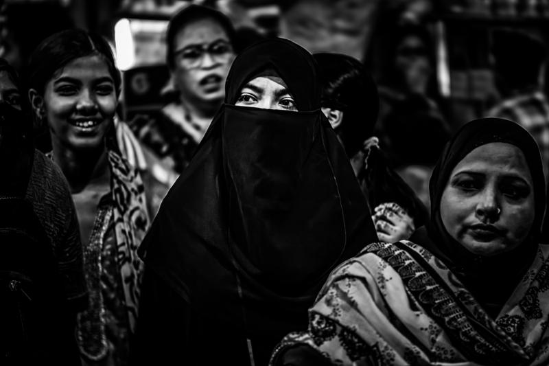 Several women turn toward the camera, their faces and patterned shawls layered tightly against the darkened market backdrop under the shop awning.