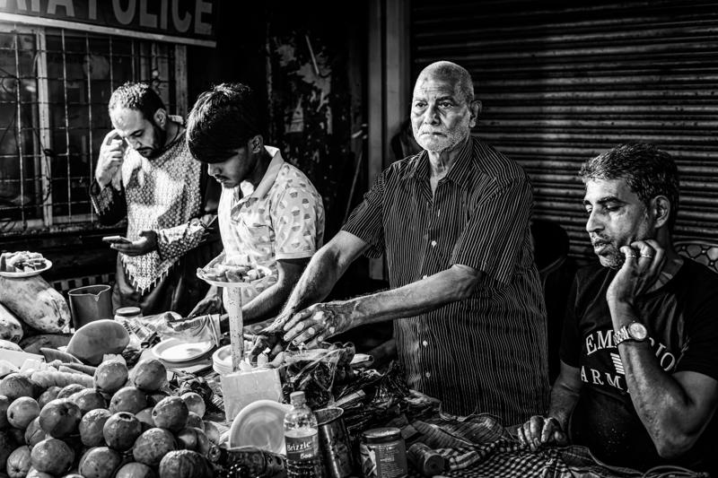 The older seller faces outward while younger men sort fruit around him, each figure caught in the cramped counter’s warm light.