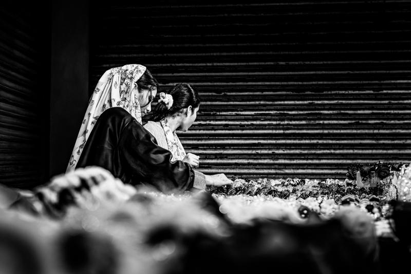 A veiled woman bends over on stone steps, her patterned shawl falling forward as visitors move through the shaded passage behind her.