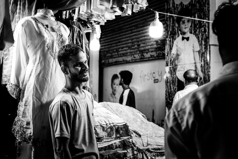 Shoppers pass the stall as a worker looks up from the counter, the hanging goods and narrow doorway crowding the monochrome frame.