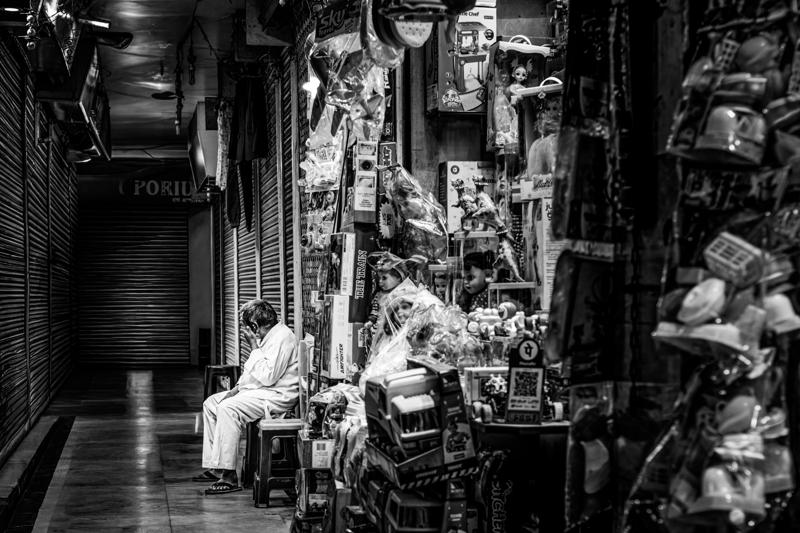 Several men sit and stand in a narrow passage lined with hanging clothes, stacked goods and small shopfronts, framing the market lane.