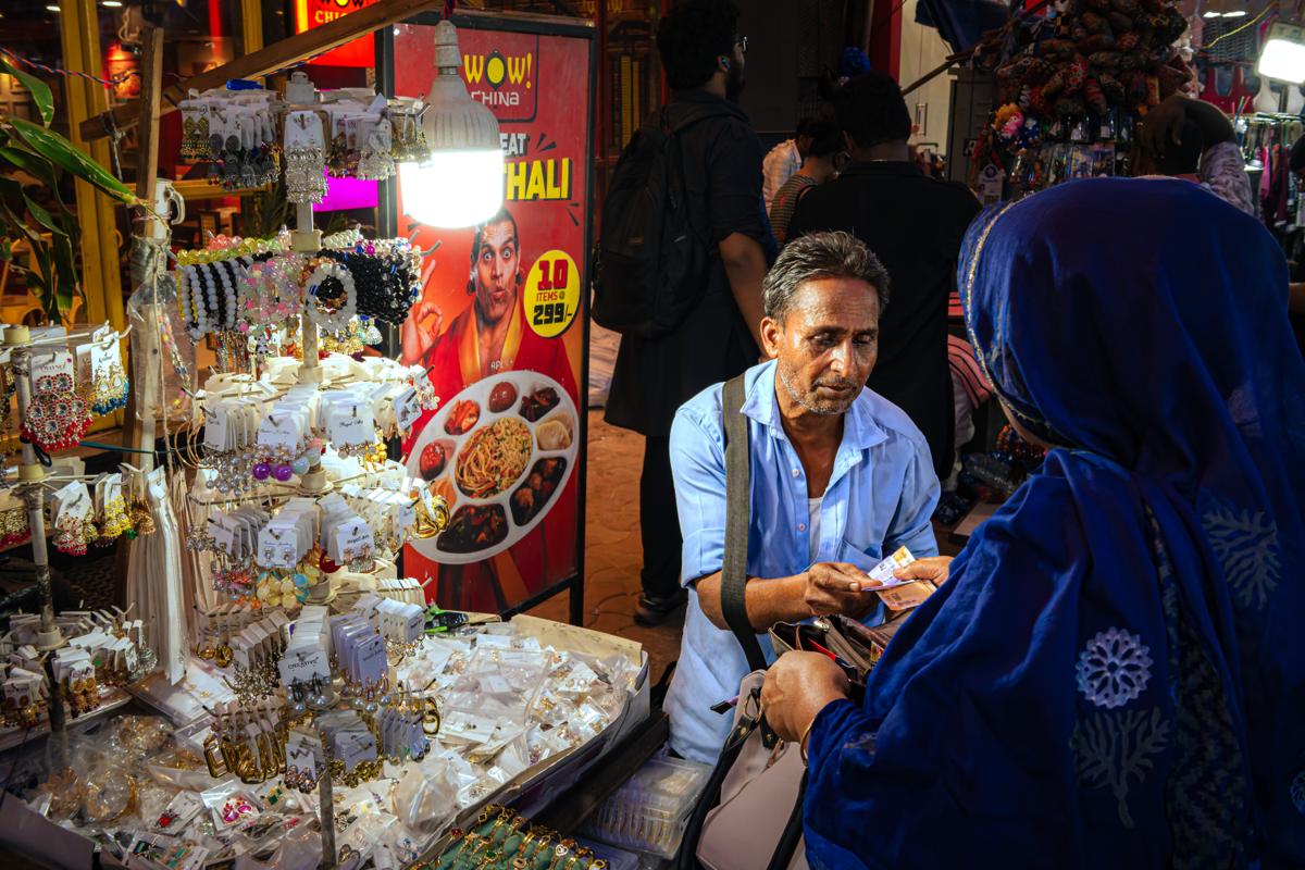A seated vendor looks out from shelves of sweets and small goods, framed by a shopper’s blue shoulder and bright packets overhead.