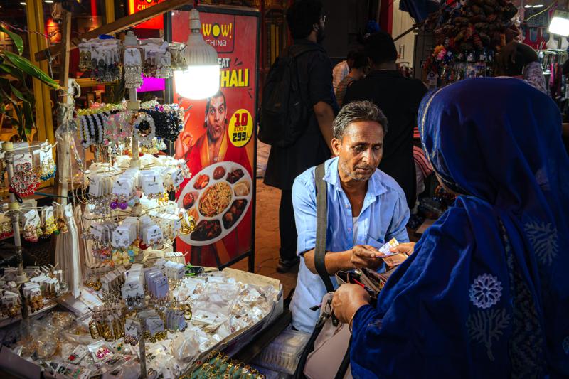 A seated vendor looks out from shelves of sweets and small goods, framed by a shopper’s blue shoulder and bright packets overhead.