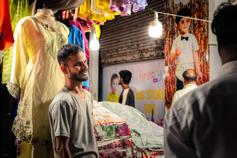 A man in a pale shirt looks across the crowded stall, framed closely by passing shoppers and a jumble of colourful goods.