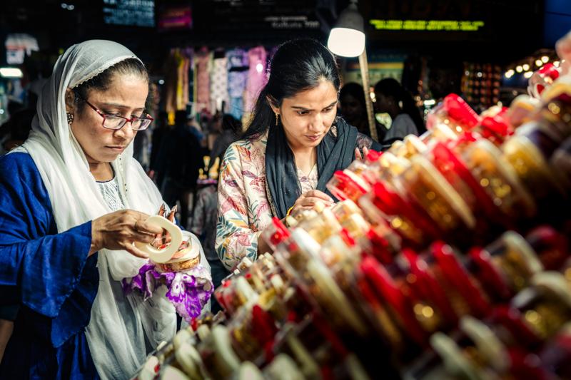 Two women examine small items beneath hanging clothes and shadowed shop bulbs, their contrasting headscarves anchoring the colourful crowd, nearby, at dusk.