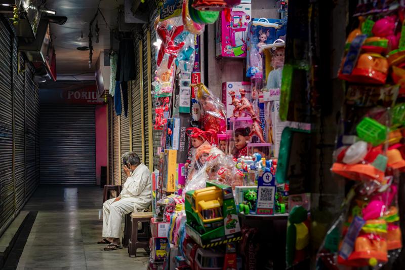 A narrow market passage glows with hanging fabric and bright packets, while a seated vendor watches from the edge of the lane.
