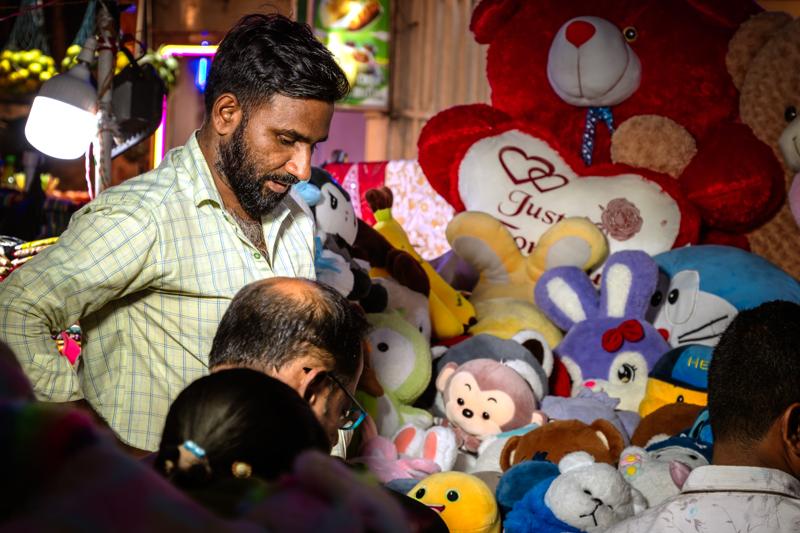 A shopkeeper leans over plush toys as a child reaches in; red hearts and crowded shelves turn the stall into a scene.