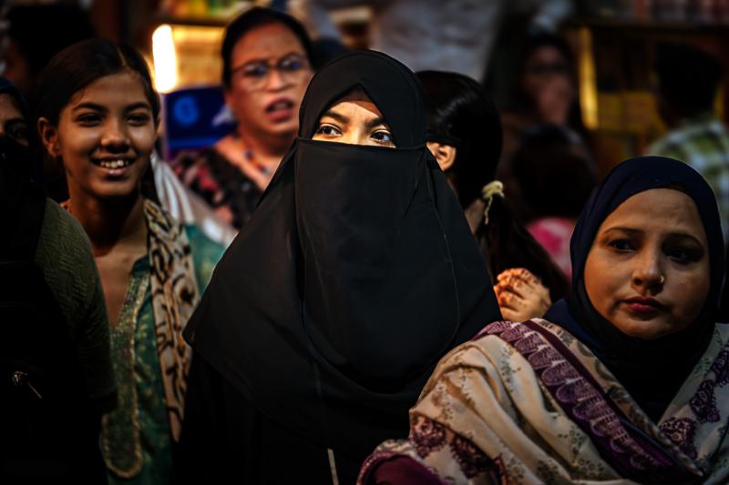 A woman in a black veil meets the camera among several shoppers, the faces and patterned shawls filling the crowded street view.