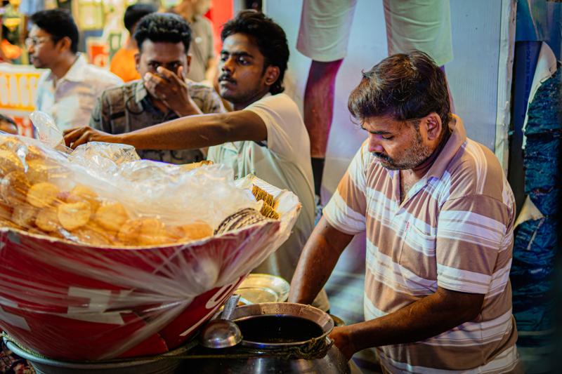 A seated vendor reaches across bowls of snacks while a customer waits nearby; warm bulbs illuminate the close, busy stall, at work.