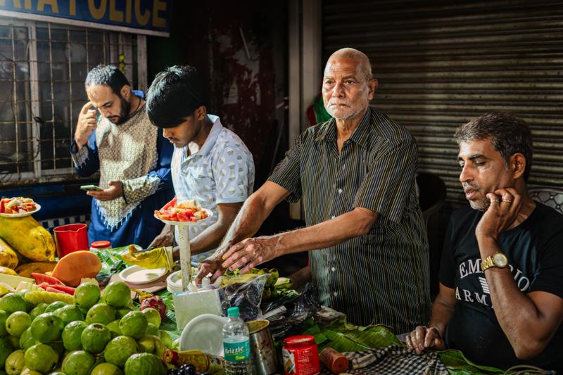 A vendor leans over his produce while two men sort fruit, their faces framed by hanging goods and the stall’s warm light.