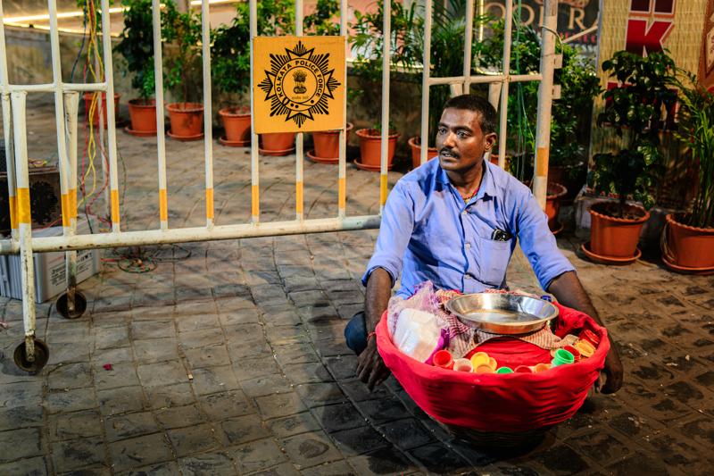 A man sits outside the lowered shutter with small goods arranged beside him, his white shirt catching the shop’s soft warm light.