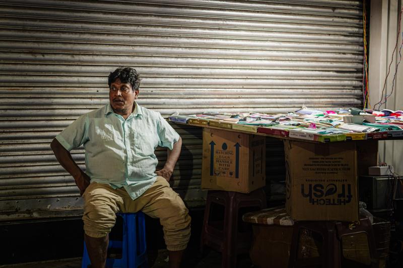 A man sits on a blue stool beside the shuttered shop, boxes and hanging wares around him under one warm bulb overhead.
