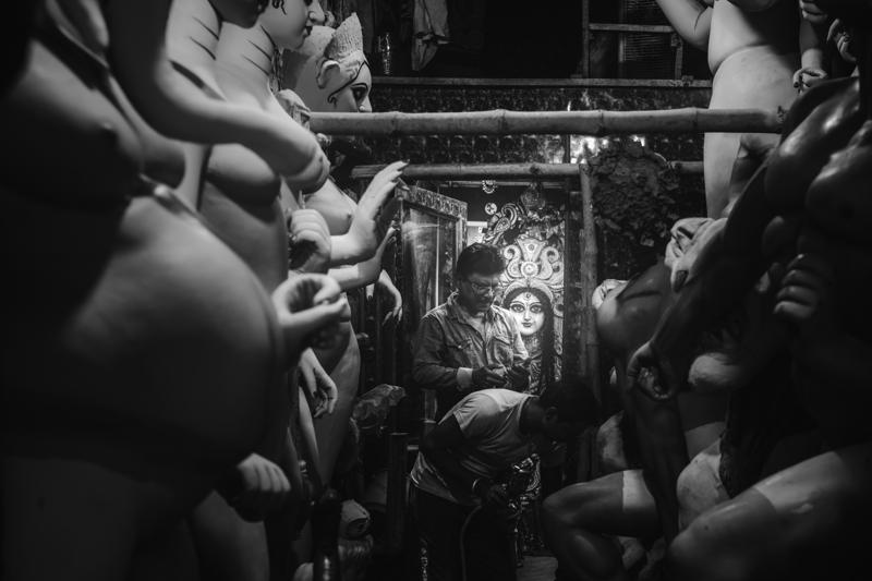 Several clay arms reach inward around a small sculpted face, creating a crowded workshop view where one bright eye holds the centre.