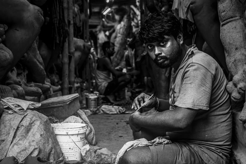 A seated artisan looks toward the lens between unfinished clay bodies, his pale shirt and focused expression emerging from the crowded studio.