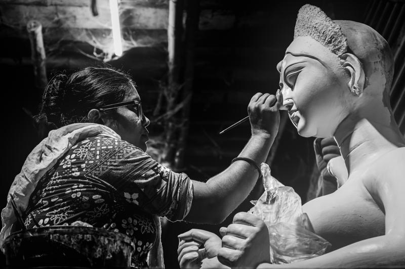 A woman leans close to a clay figure, painting its face while another worker steadies the statue inside the crowded workshop room.