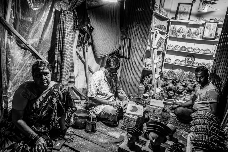 People gather along the narrow path as sunlight breaks between the buildings, turning the crowded market passage into a band of gold.