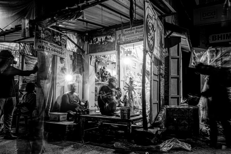 A narrow storefront glows within a dark street, revealing a bent worker and hanging goods while the surrounding façades recede.