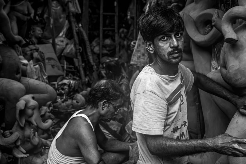 A young artisan glances toward the camera amid rows of unfinished figures, his pale shirt briefly separating him from the crowded workshop.