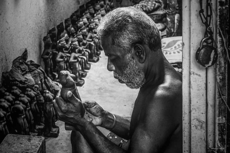 A sculptor leans over a clay figure, his hands working between pale unfinished forms as the monochrome workshop falls into soft shadow.