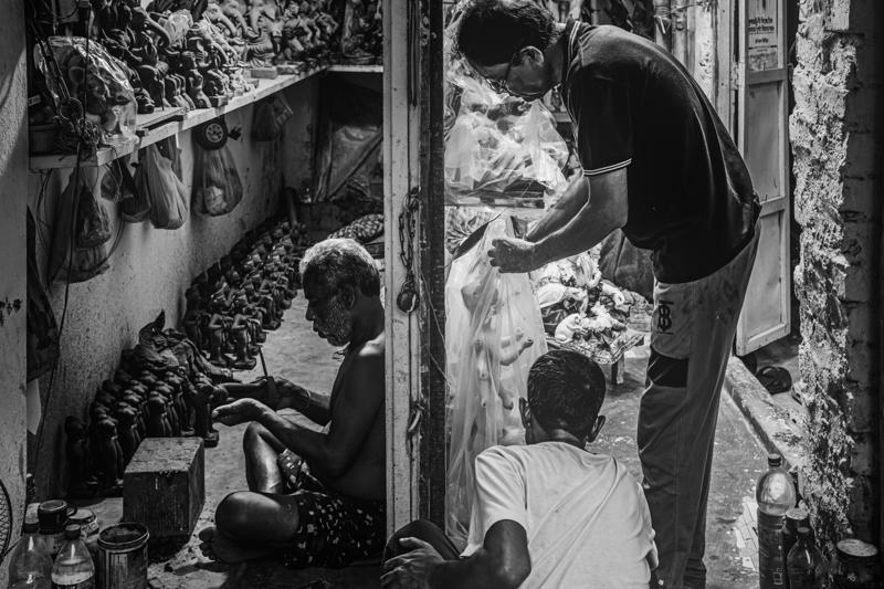 An artisan bends beside a clay figure, pale statues crowding the monochrome workshop around his bare shoulders and focused face in shadow.