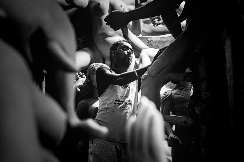 A craftsman reaches toward an painted idol’s cheek as another worker watches, the unfinished face and layered hands sharing the close frame.