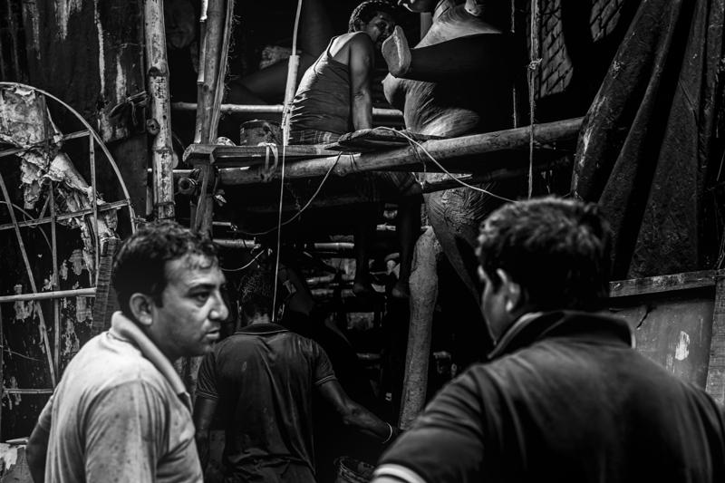 A man works at a narrow counter surrounded by sculpted faces and tools, the lamp revealing only part of the busy room.