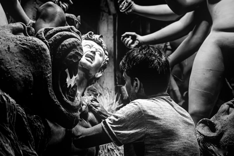 An artisan reaches toward a sculpted face as others work close by, their hands, tools and unfinished forms filling the crowded workshop.