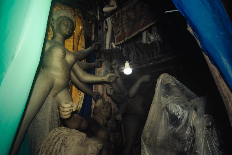An artisan works among covered clay figures inside the crowded studio, with the blue-lit opening drawing the eye through the narrow space.
