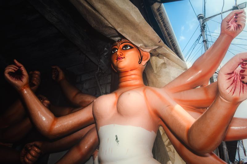 A large idol’s pink face tilts upward beneath draped fabric, its bright eyes and beaded crown emerging from the close, shadowed pandal.
