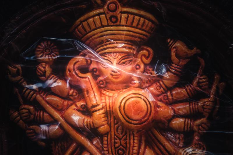 Warm light reveals the goddess’s painted eyes and beaded crown, with overlapping figures and textured decoration surrounding her face, nearby, up close.