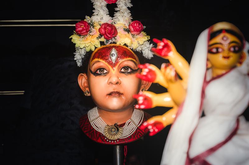 A child in bright costume looks straight into the camera, with flowered headwear and another small figure soft behind her.