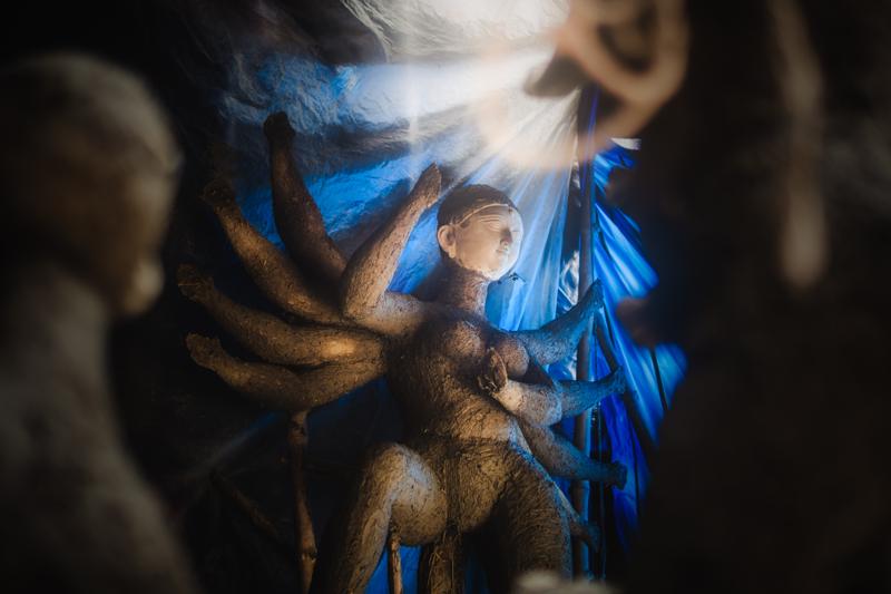 A painted face appears through a sweep of blue fabric, the blur making the goddess seem to emerge from the workshop shadows.